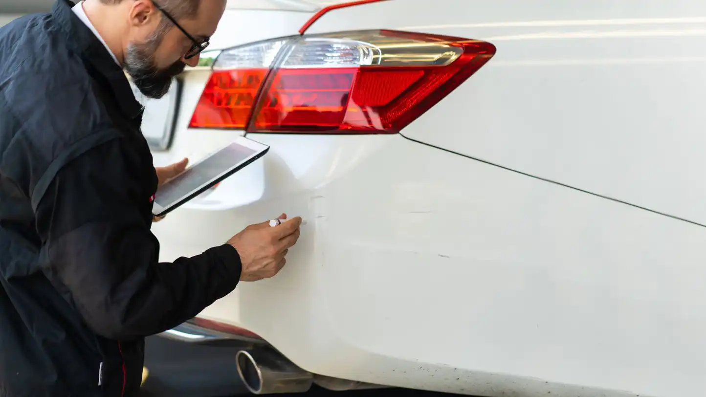 Techniker hält ein Tablet in der Hand, während er ein weißes Auto auf kosmetische Schäden untersucht.