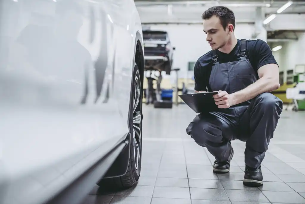 Technician holding a clipboard while inspecting a white car in repair shop.