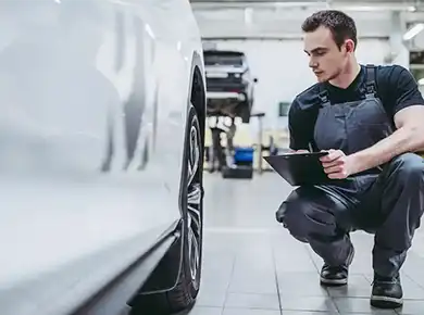 Technician holding a clipboard while inspecting a white car in repair shop.