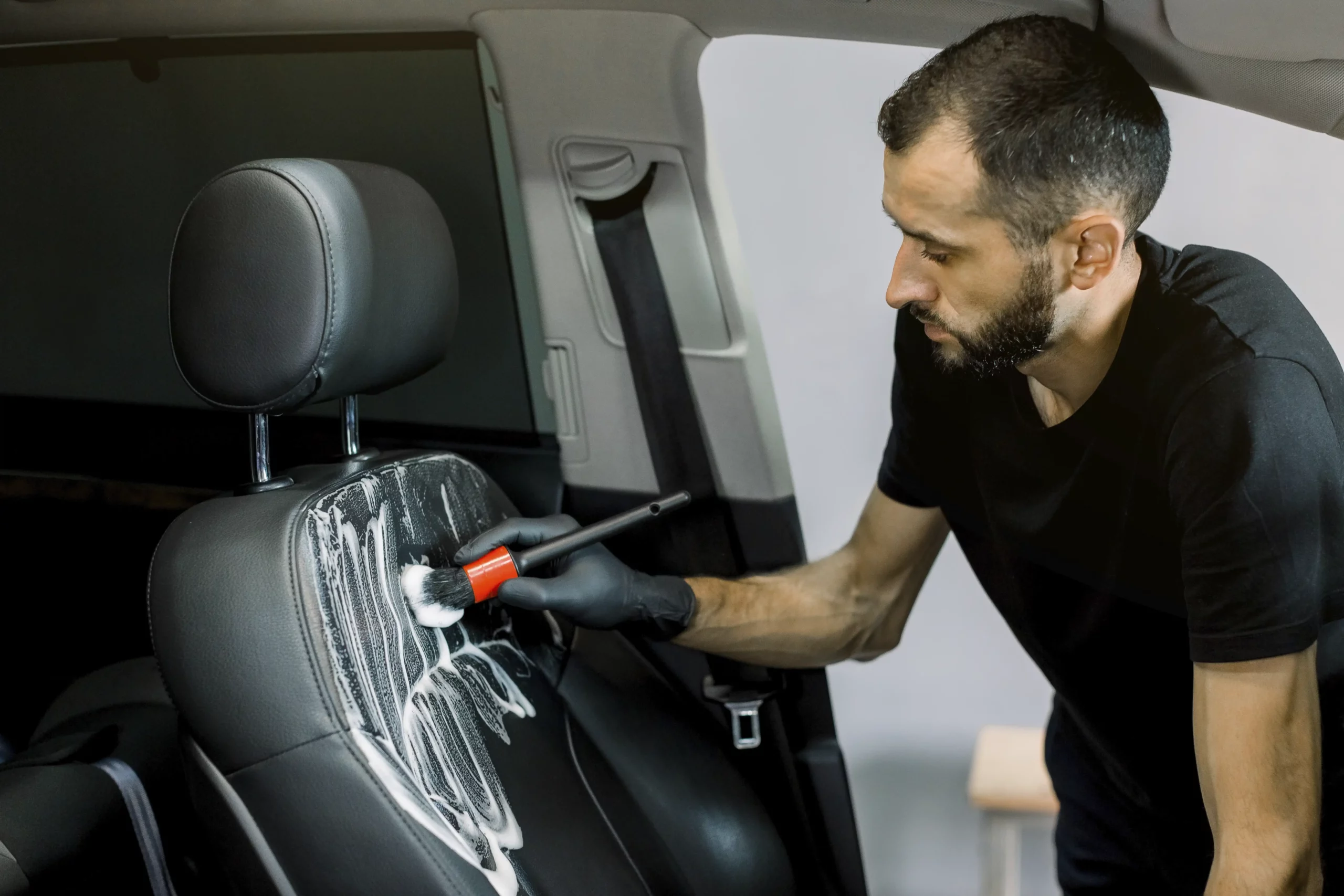 The technician is cleaning a black leather car seat with a detailing brush.